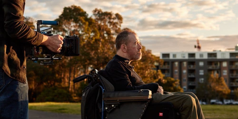 A man using a wheelchair being filmed outdoors.